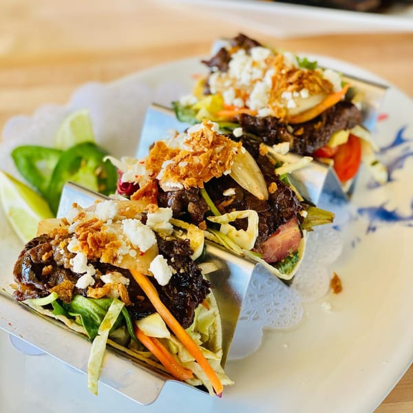 Three Asian fusion tacos with meat, vegetables, cheese, and crispy toppings, served with lime and jalapeños.