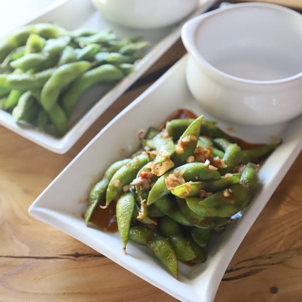 Two white rectangular dishes of edamame, one plain and one with garlic sauce, on a wooden table.