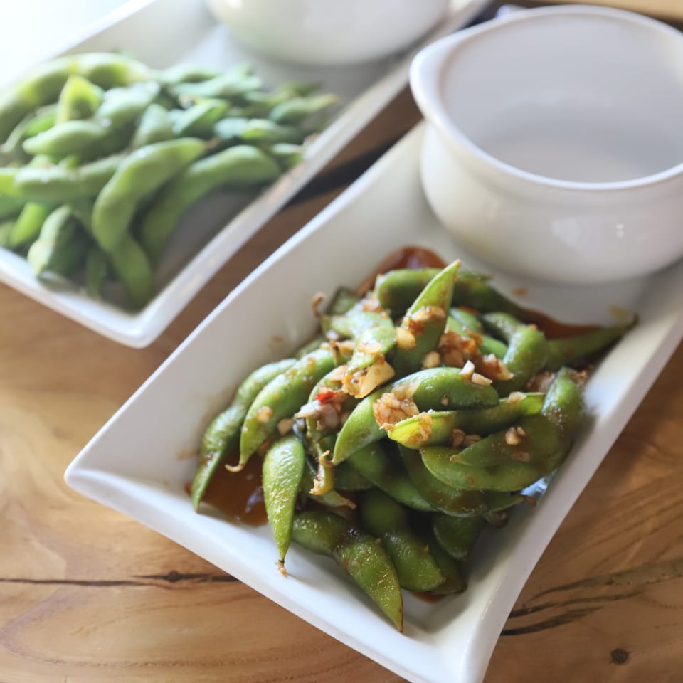 Two white rectangular dishes of edamame, one plain and one with garlic sauce, on a wooden table.
