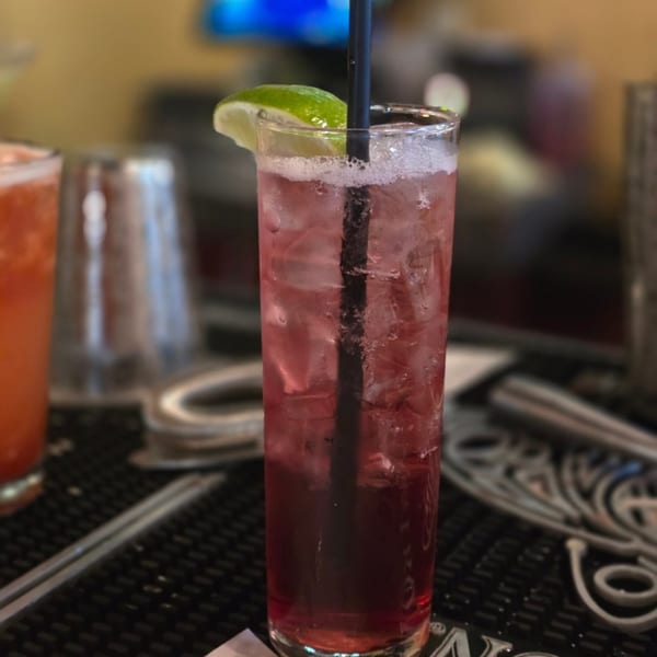 A tall glass of red iced drink with a lime wedge and black straw on a bar.