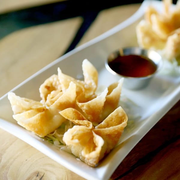 Crispy golden crab rangoons with a side of dark dipping sauce on a white rectangular plate.